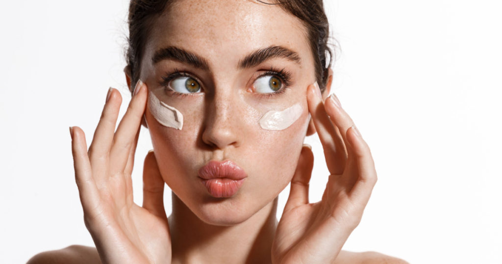 Close up beauty girl with freckles and thick eyebrows, applying moisturizing skincare cream, lotion or mask for skin lifting and anti-aging detoxifying effect, white background.
