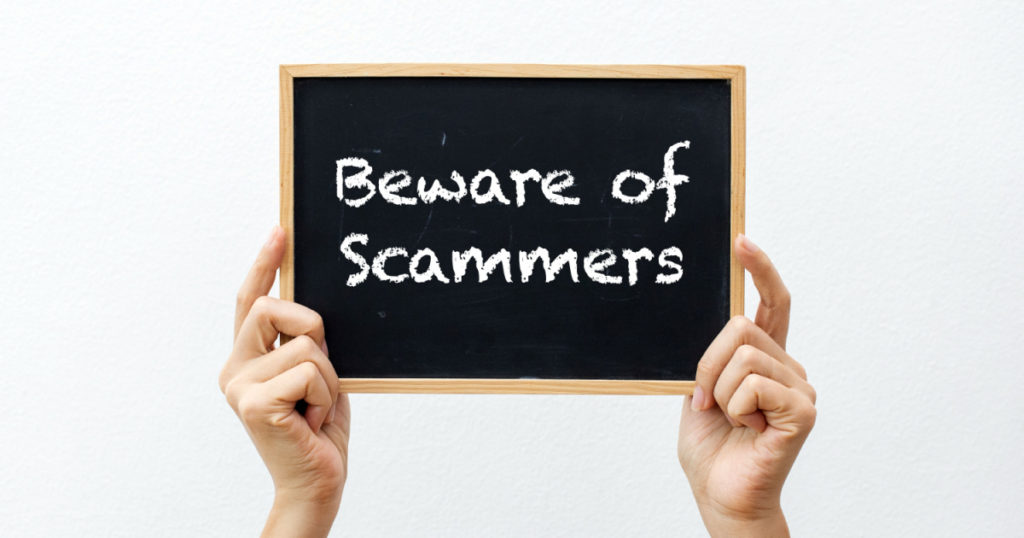 Cropped hands holding a chalkboard with "Beware of Scammers" against a white background

