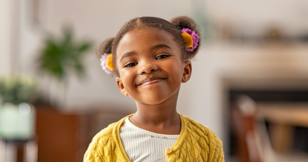 Smiling cute little african american girl with two pony tails looking at camera. Portrait of happy female child at home. Smiling face a of black 4 year old girl looking at camera with afro puff hair.
