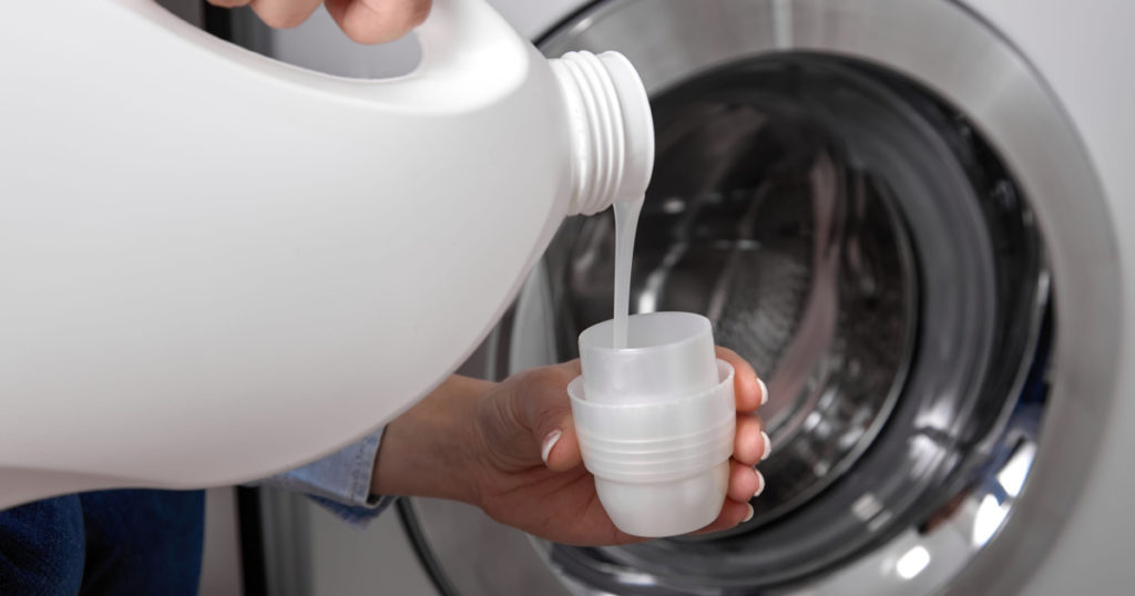 Against background of drum of steel-colored washing machine, woman pours liquid washing gel into plastic cap. A girl in a white T-shirt carefully pours a transparent conditioner for flattening laundry
