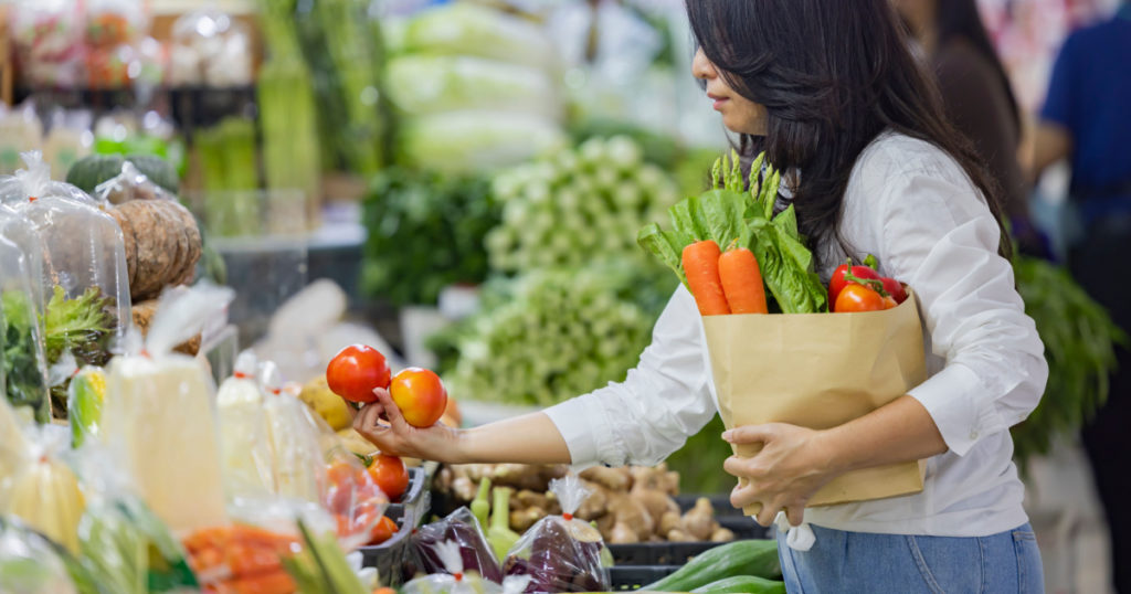 Asian woman holding large paper shopping bag of food. Woman choosing food products in grocery shop or supermarket holding vegetables in hand and thinking what to cook.
