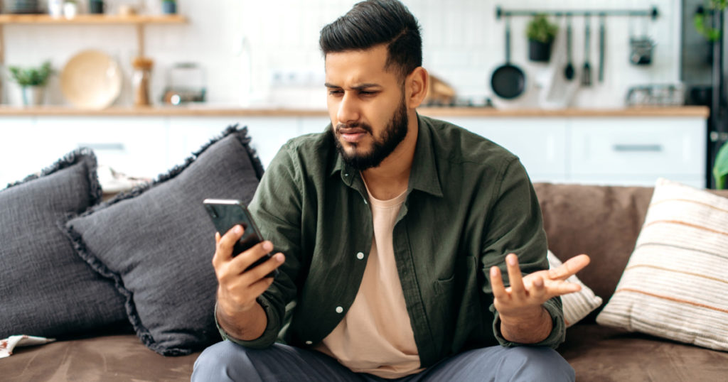 Disappointment puzzled mixed race young man in casual wear, sits on sofa in living room, using mobile phone, texting online, browsing social networks, reads news, looks at screen in confusion
