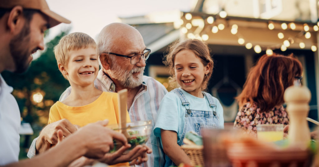 Happy Senior Grandfather Talking and Having Fun with His Grandchildren, Holding Them on Lap at a Outdoors Dinner with Food and Drinks. Adults at a Garden Party Together with Kids.
