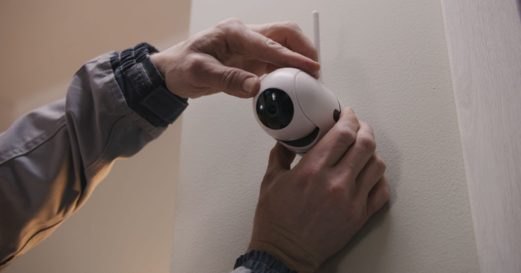 Installer in uniform puts security camera on wall fastening and connects it to system with cable. Man installs cameras in house. Concept of CCTV cameras, monitoring, safety and privacy.
