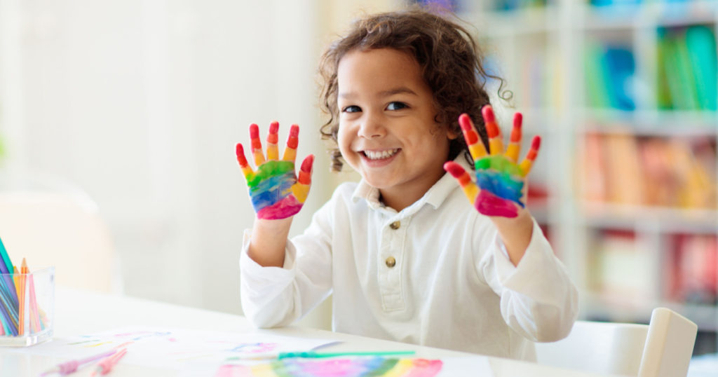 Child drawing rainbow. Paint on hands. Remote learning and online school art homework from home. Arts and crafts for kids. Little boy drawing bright picture. Creative kid playing and studying.
