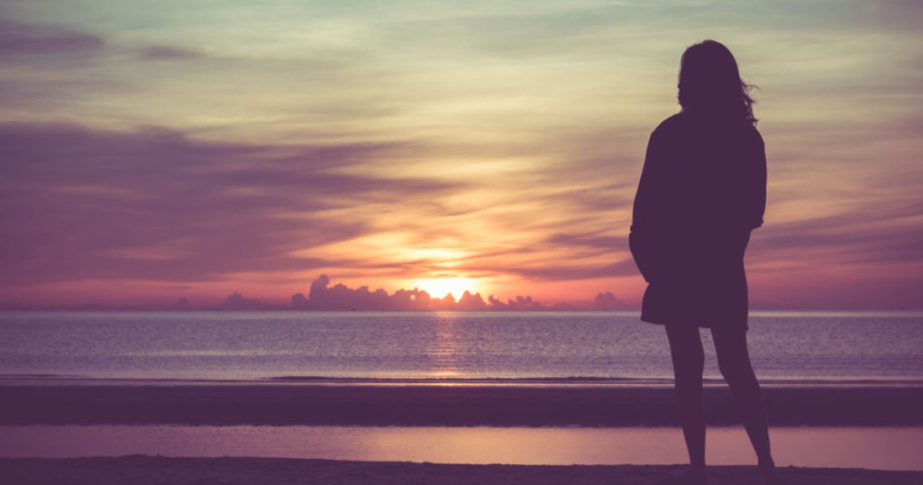 Young woman standing on the beach in sea and looking to sunrise.
