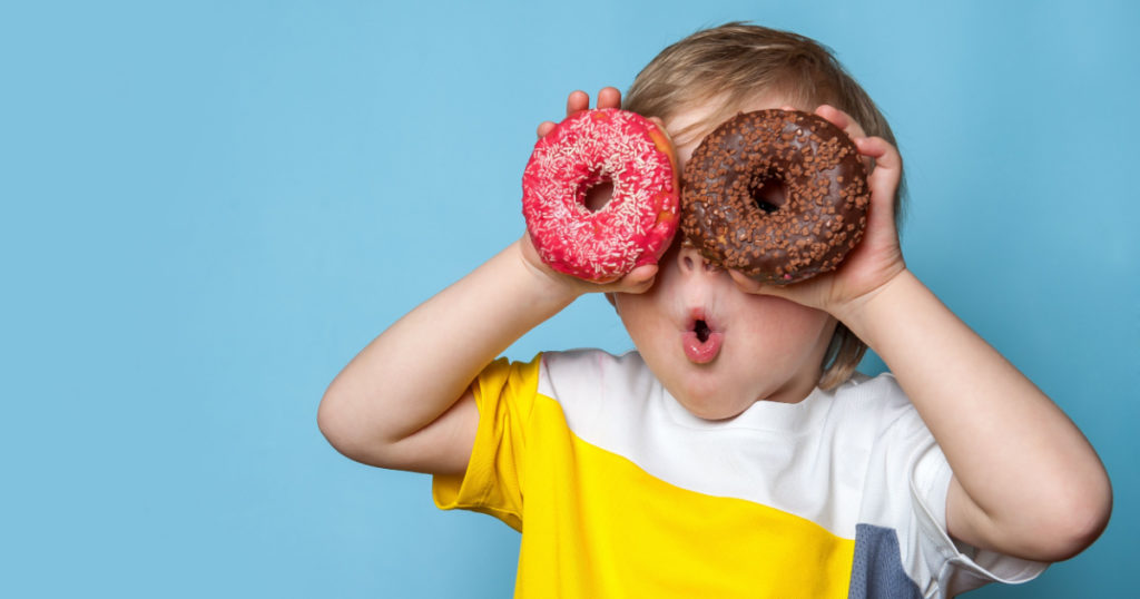 Little happy cute boy is eating donut on blue background wall. child is having fun with donut. Tasty food for kids. Funny time at home with sweet food. Bright kid.
