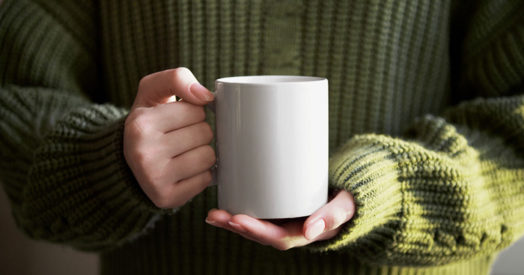 female hand holding white mug with blank copy space for your advertising text message or promotional content, sweet coffee or tea. Girl in green sweater holding white porcelain mug mock up