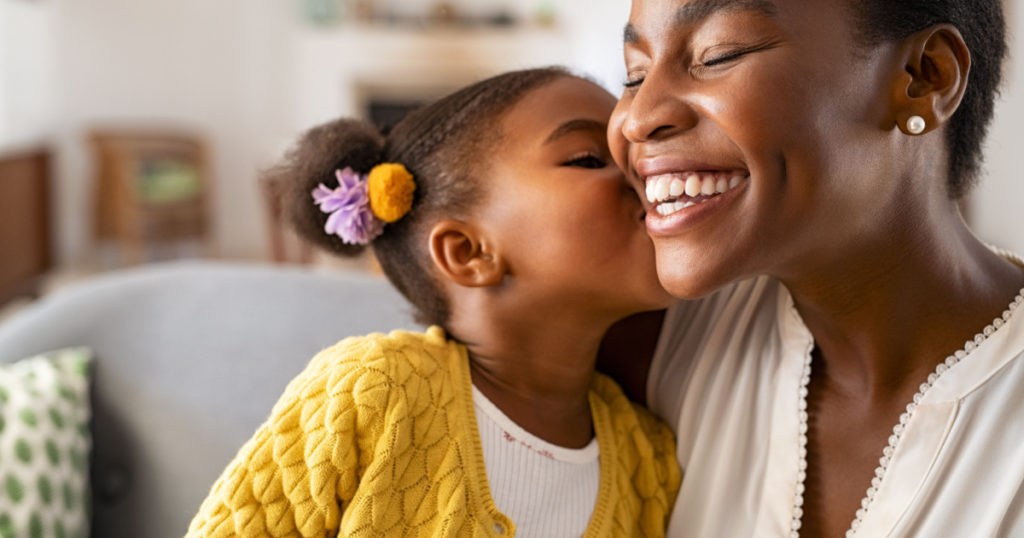 Close up of beautiful daughter kissing mother on cheek at home. African little girl giving kiss to happy mother. Lovely black female child kissing cheerful and proud woman on cheek for mother's day.
