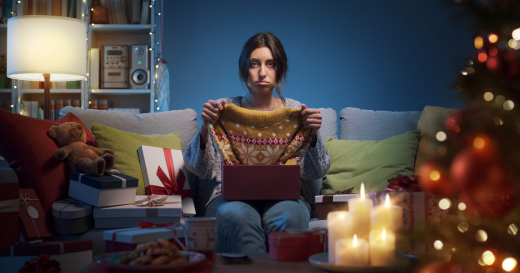 Sad disappointed woman opening Christmas gifts at home, she has received an ugly Christmas sweater