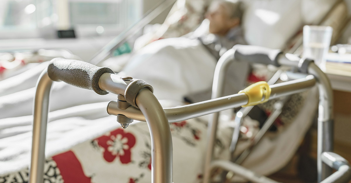 patient in hospital bed out of focus in background, with metal walking aid in focus in the foreground