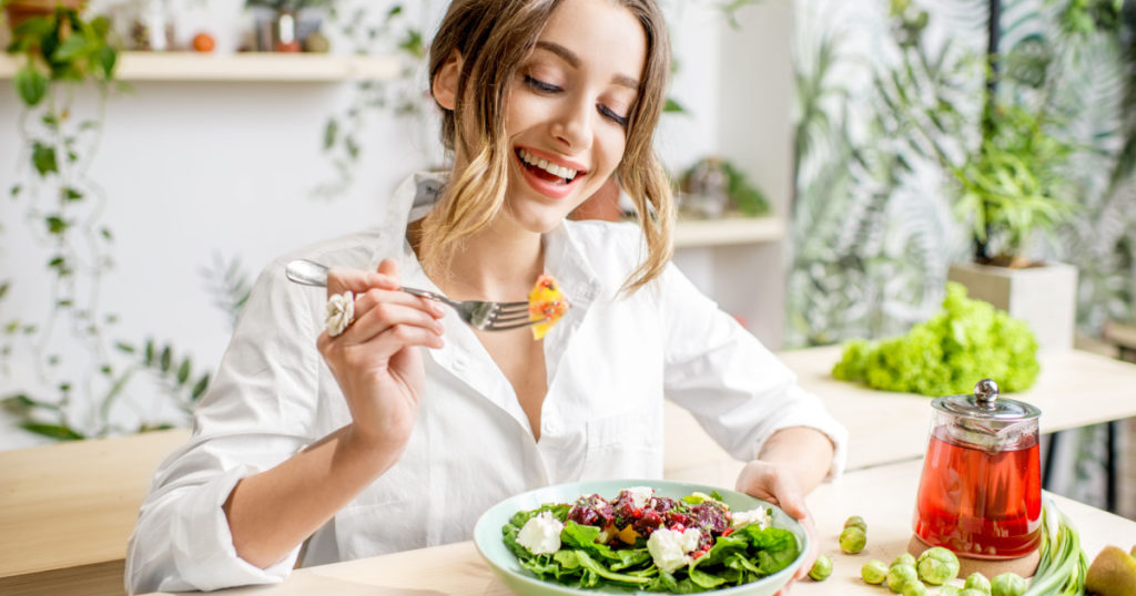 Young woman eating healthy food sitting in the beautiful interior with green flowers on the background
