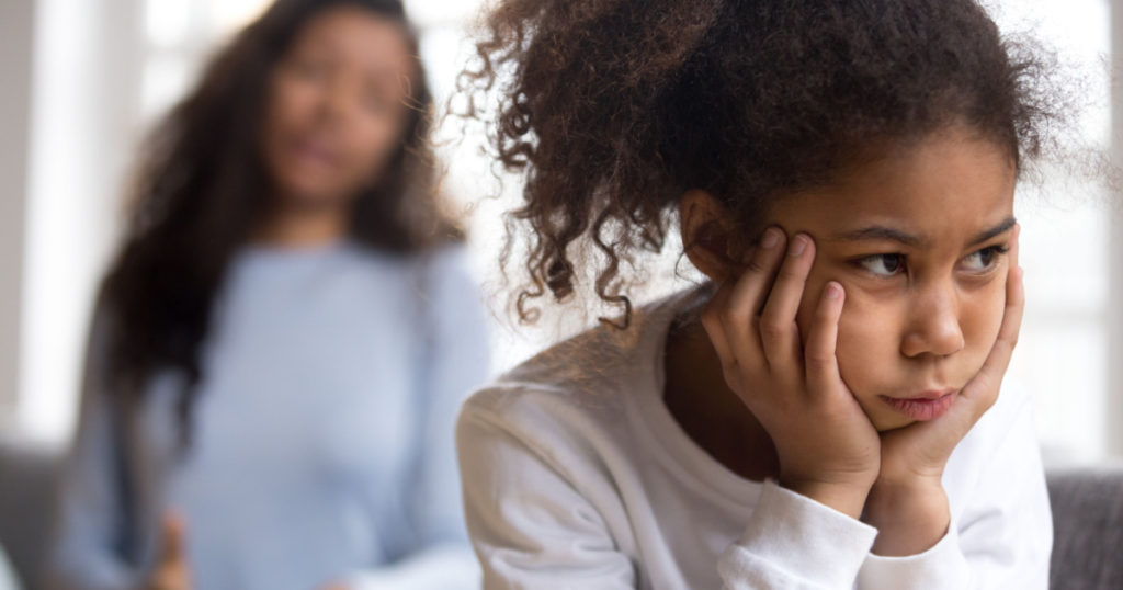 Offended African American preschooler girl looking in distance, holding head in hands, talking mother on background, family conflict, child punishment, complicated relationships with parent, close up
