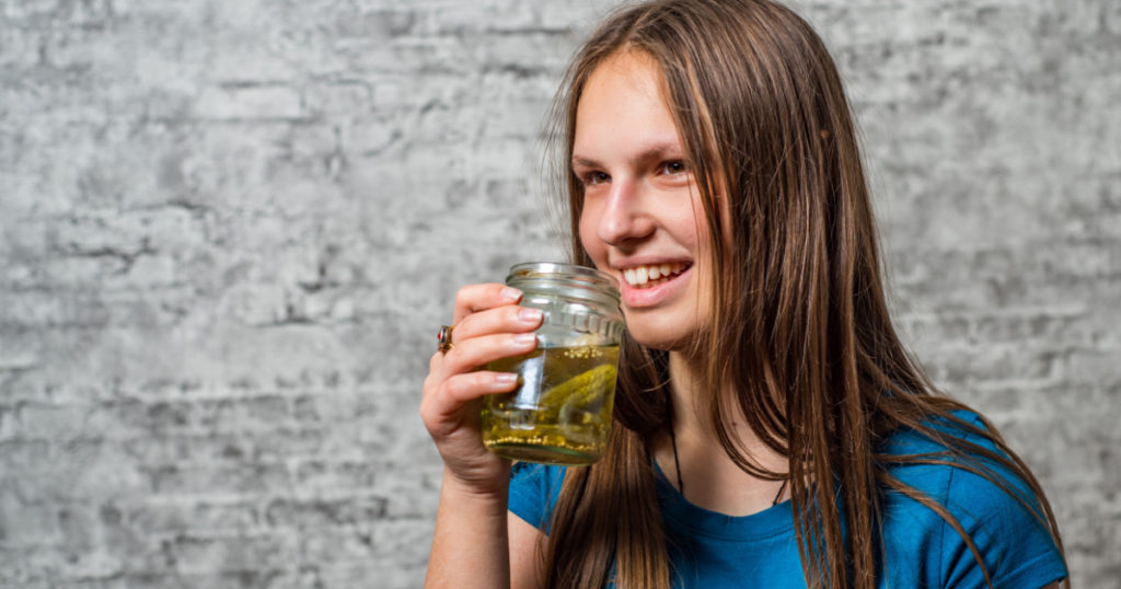 portrait of young teenager brunette girl with long hair drink Pickle juice in can of pickled cucumbers on gray wall background
