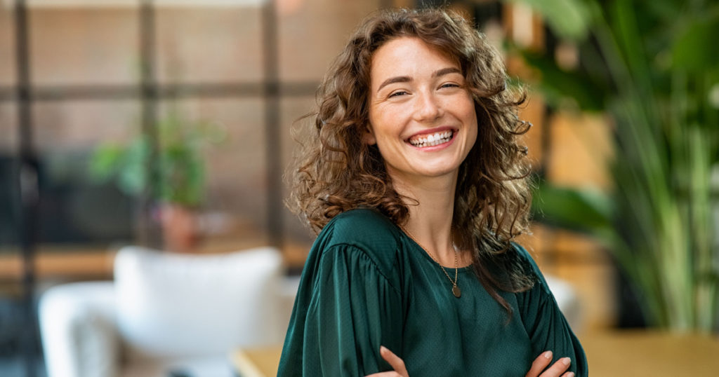 Portrait of young smiling woman looking at camera with crossed arms. Happy girl standing in creative office. Successful businesswoman standing in office with copy space.