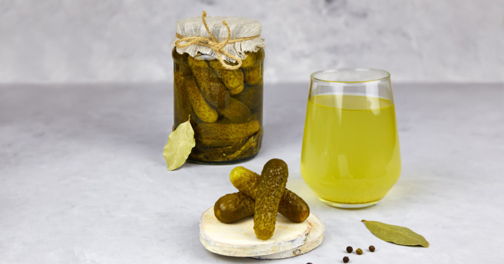 Pickled cucumbers in a glass jar and pickle juice in glass on a light background. Homemade fermented or marinated cucumbers. Healthy energy drink for athletes.
