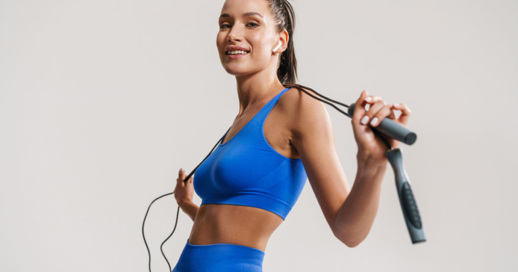 Young white fitness woman in sportswear with ponytail standing over white wall background, holding skipping rope