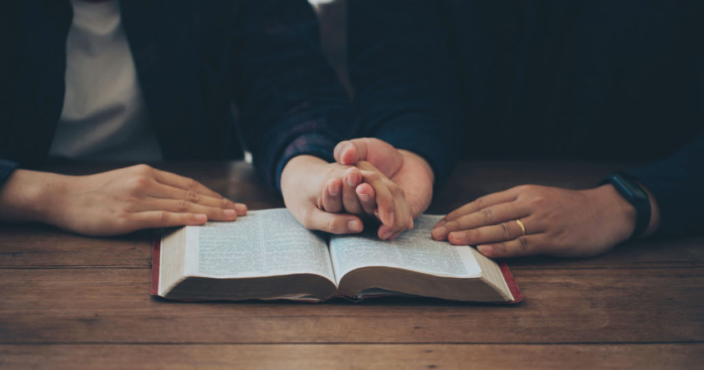 Two Christian couples holding each other's hands praying together over the bible on a wooden table. Begging for forgiveness and believing in goodness. Christian life crisis prayer to god.