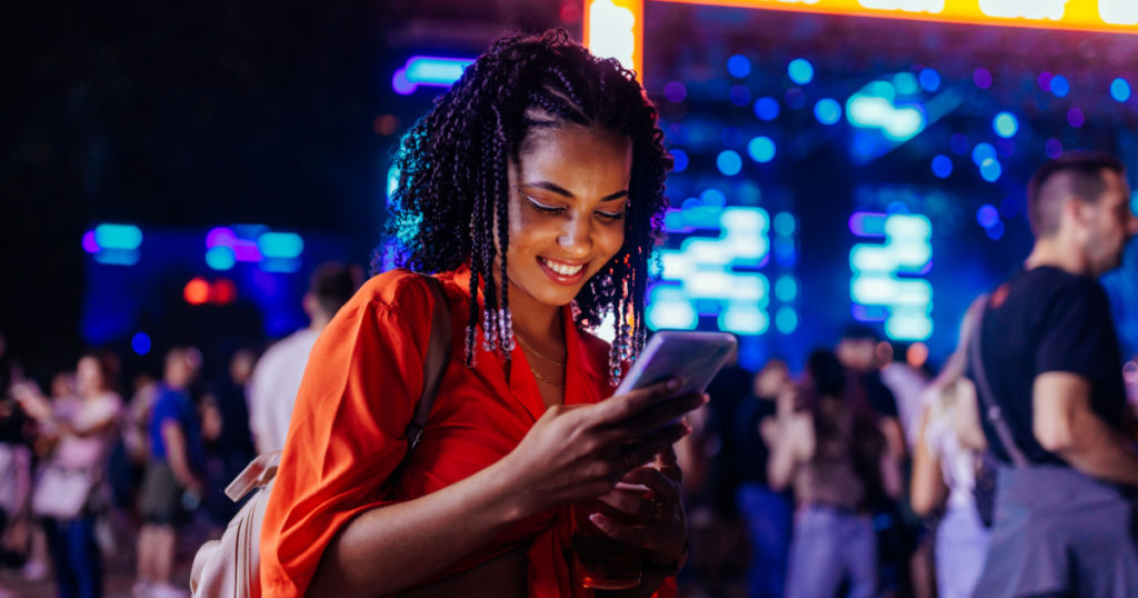 A young African American girl is with a smartphone at a music festival, texting with her friends.