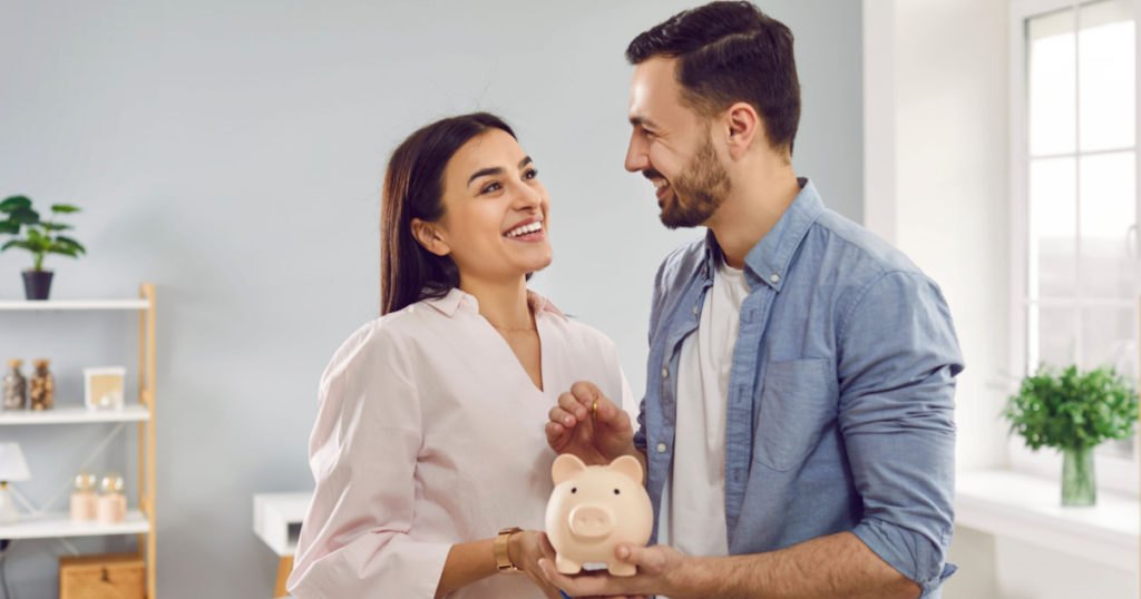 Smiling, happy young family together put coins in piggy bank to save money. Married couple are planning to save up finances. Savings, investments, financial freedom, business, hope for success.