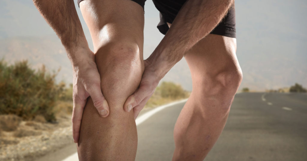 young sport man with strong athletic legs holding knee with his hands in pain after suffering muscle injury during a running workout training in asphalt road in muscular or ligament wound
