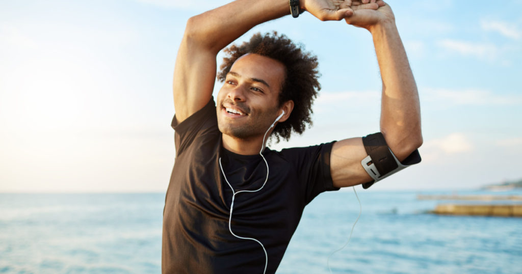 Portrait of a smiling afro-american sports man stretching his muscular arms before workout by the sea, using music app on his smartphone. Dark-skinned athlete warming up before running.
