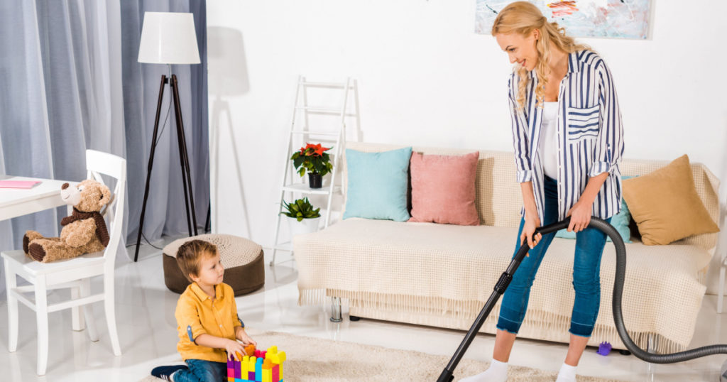 cute little boy playing with colorful blocks and looking at pregnant mother cleaning carpet with vacuum cleaner
