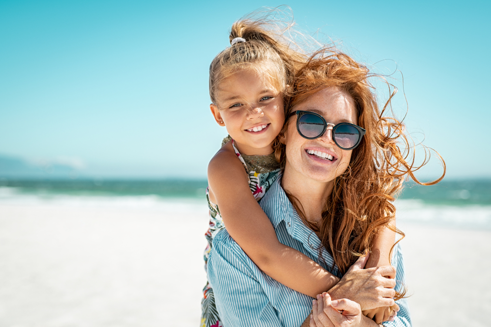 Smiling mother and beautiful daughter having fun on the beach. Portrait of happy woman giving a piggyback ride to cute little girl with copy space. Portrait of kid embracing her mom during summer.