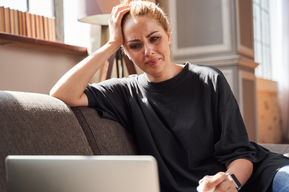 Upset young woman crying and putting her head on the hand while looking at the laptop screen at her psychologist