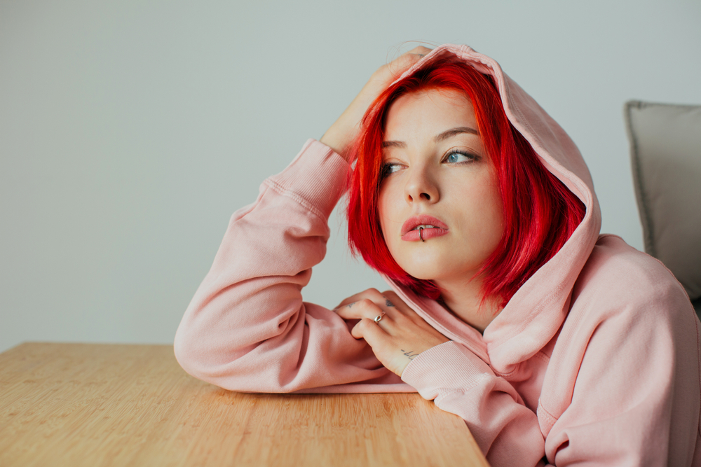 Portrait of a cool female with bright red hair, lip piercing and hoodie holding head on desk looking out the window