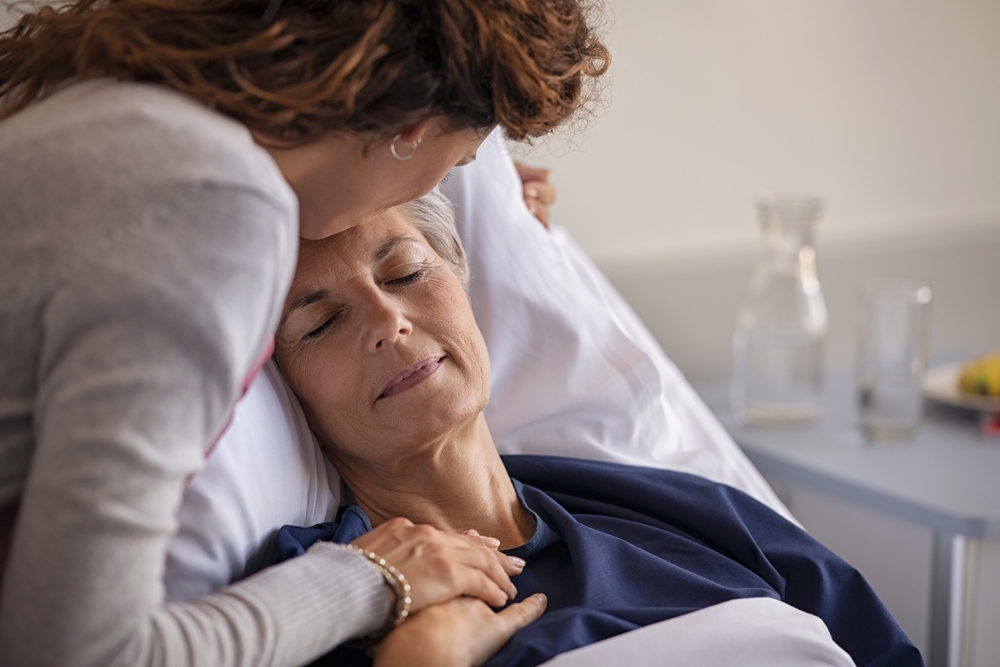 Close up of lovely daughter visiting and kissing sick mother on forehead while resting in hospital bed. Senior patient laying on gurney with eyes closed while daughter assuring a speedy recovery.