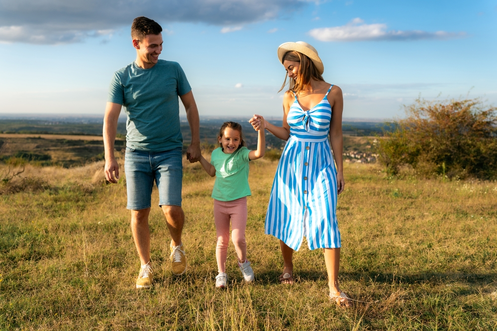 Mother and father are holding their daughter's hands as they walk through the field, having fun and laughing