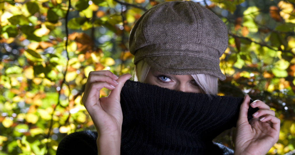 Mysterious young blond woman standing peering at the camera over the top of the raised cowl neck collar of her sweater against colorful autumn or fall leaves