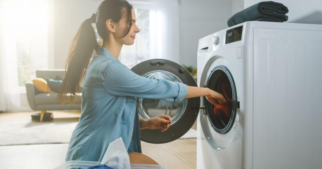 Beautiful Smiling Brunette Young Woman Sits in Front of a Washing Machine in Homely Jeans Clothes. She Loads the Washer with Dirty Laundry. Bright and Spacious Living Room with Modern Interior.