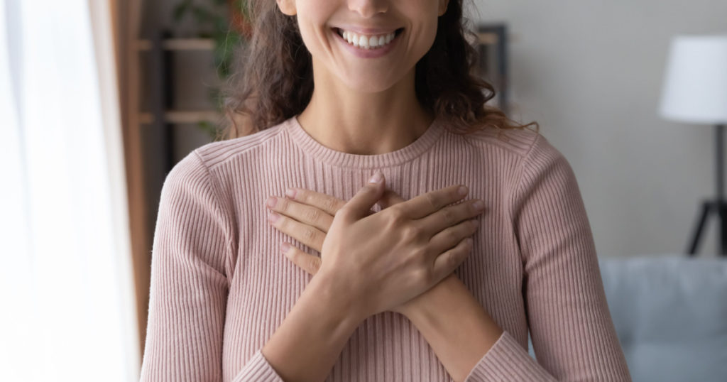 Close up focus on happy sincere female holding folded hands on chest. Emotional positive kind candid millennial woman feeling thankful indoors, showing gratitude sign, believe faith charity concept.
