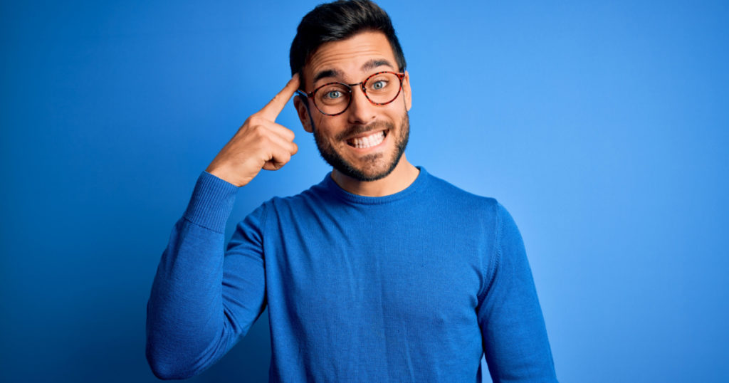 Young handsome man with beard wearing casual sweater and glasses over blue background Smiling pointing to head with one finger, great idea or thought, good memory
