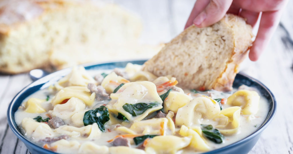 Fresh Tortellini Soup with Italian sausage, spinach and carrots. hand dipping homemade artisan bread. Selective focus on pasta with blurred background.