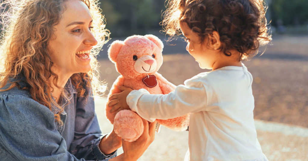 Curly single woman giving a teddy bear to her little daughter outdoors in the park - family lifestyle concept