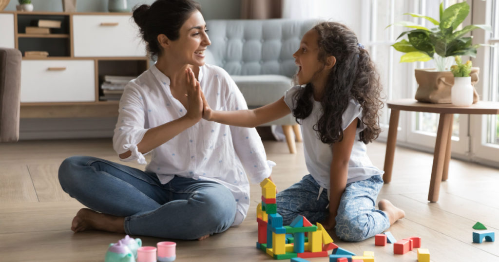 Happy beautiful Indian mom and pretty little daughter girl clapping hands, giving high five over wooden construction blocks, toy tower, castle on heating floor, celebrating model completing