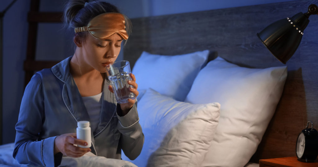 Young woman with pill bottle and glass of water in bedroom at night