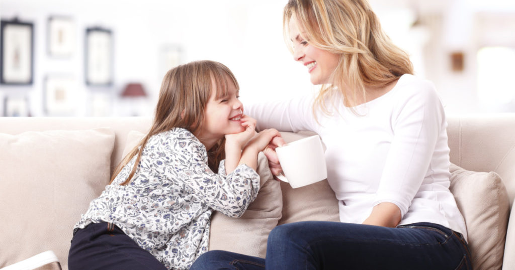 Portrait of a beautiful mother and her little girl sitting at home and sharing a happy moment together. Blond mom holding in her hand a cup of tea.