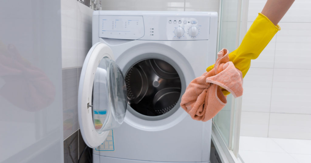Front view of unidentifiable yellow rubber gloved hand holding dirty pink towel in front of small front loading washing machine