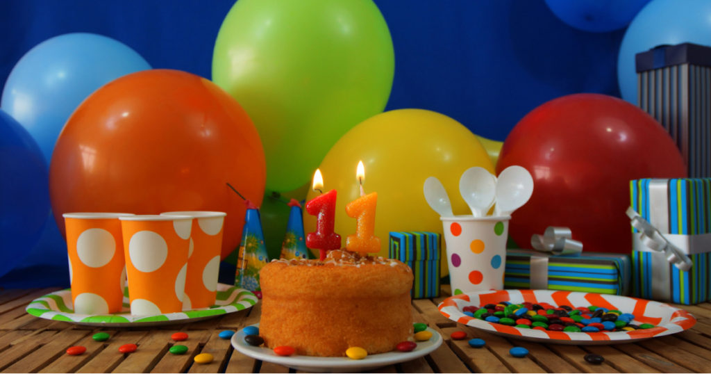 Birthday cake on rustic wooden table with background of colorful balloons, gifts, plastic cups and plastic plate with candies and blue wall in the background