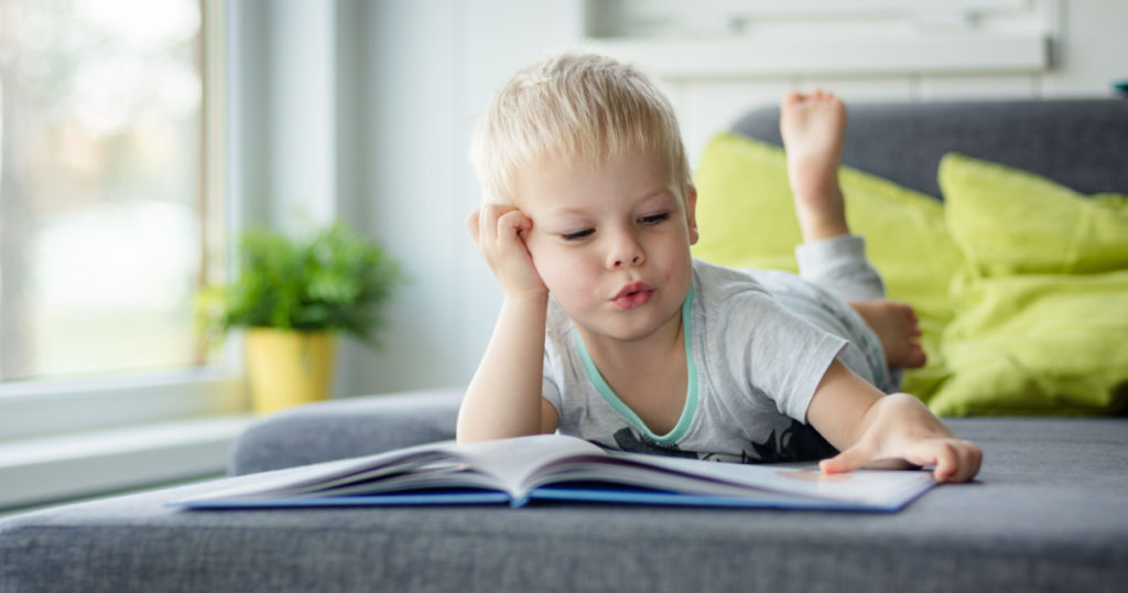Little, cheerful, three years old boy lying on his belly on the sofa, with open book in front of him.