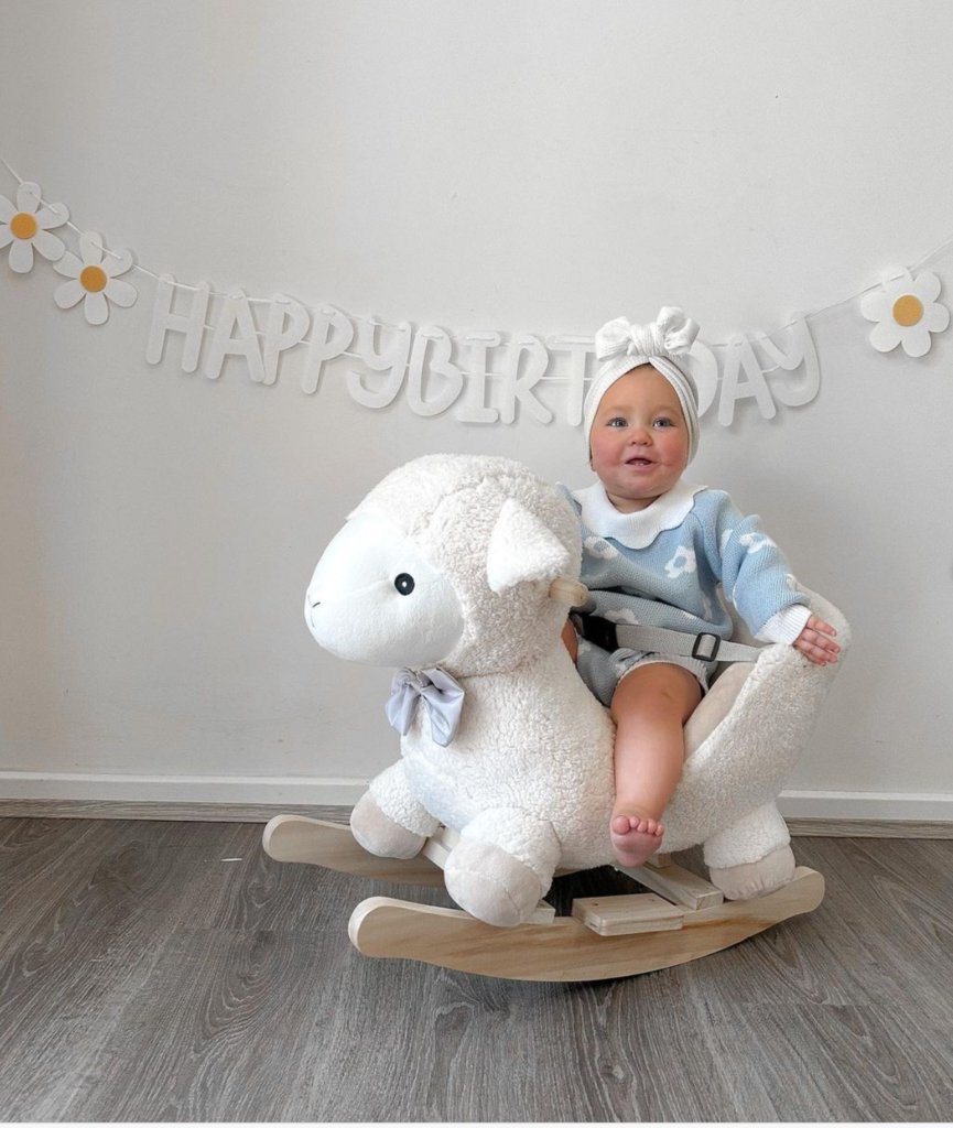 Baby sitting on a rocking sheep with happy birthday banner in the background. 