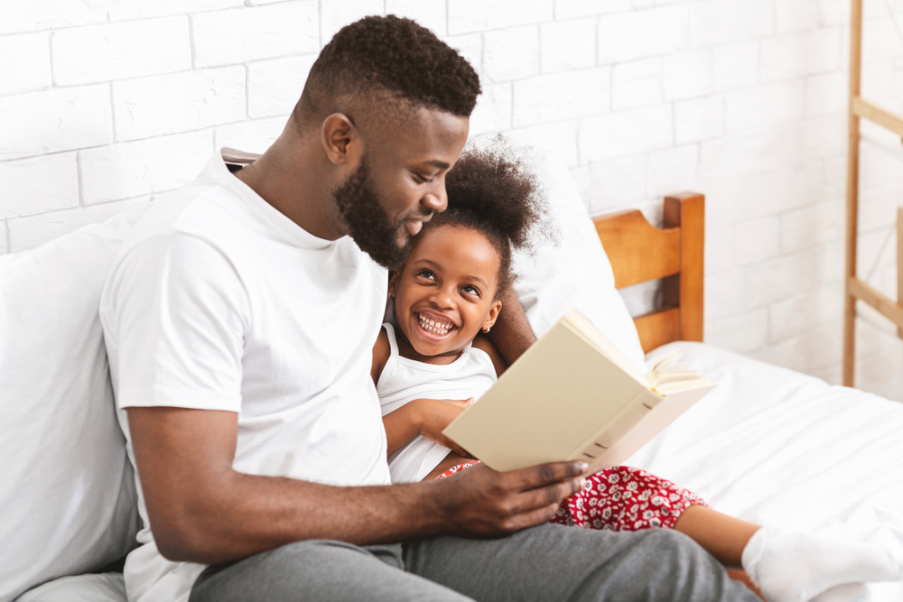 Happy evening. African american man reading fairy tale to his little daughter, sitting in bedroom, empty space