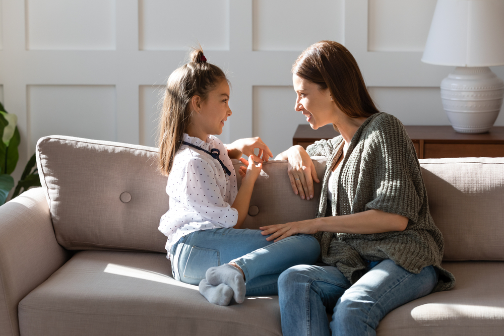Caring mother and little daughter chatting, sitting on cozy couch in living room, pretty girl sharing secrets with loving mum, having pleasant conversation, good trusted family relationship