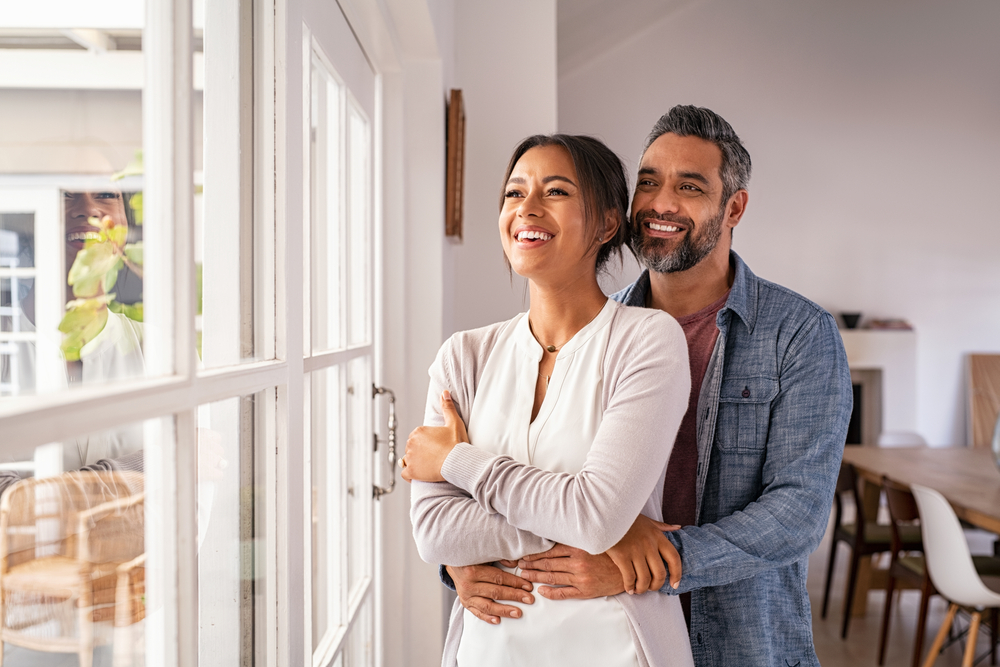 Smiling mid adult couple hugging each other and standing near window while looking outside. Happy and romantic mature man embracing hispanic wife from behind while standing at home with copy space.