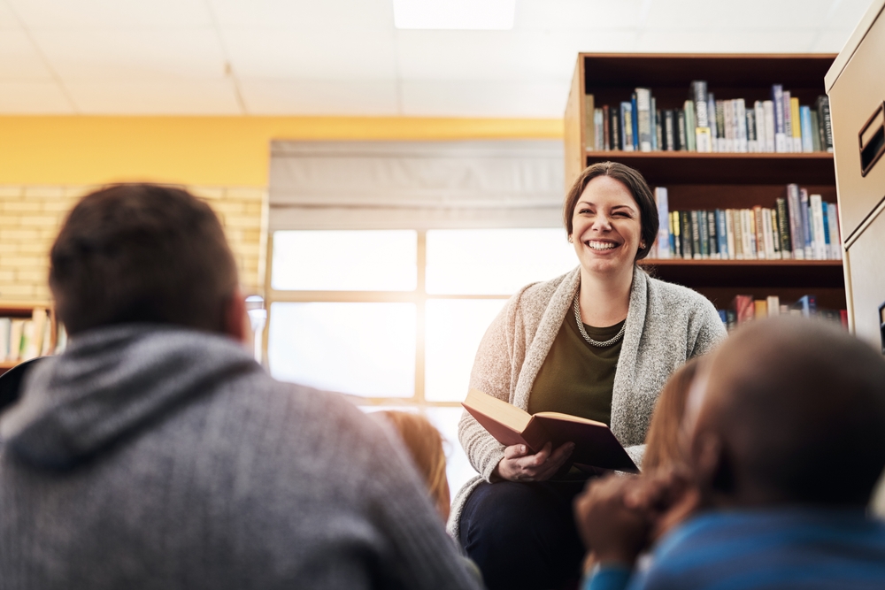 Library, kids and teacher reading book, education and learning with knowledge, storytelling and kindergarten. Educator, woman and children on floor, fun and fantasy stories with development or relax
