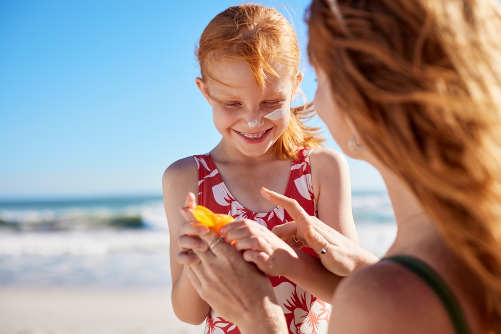 Young mother applying protective sunscreen on daughter nose at beach with copy space. Woman hand putting sun lotion on female child face at sea. Cute little girl with freckles applying sunblock cream.